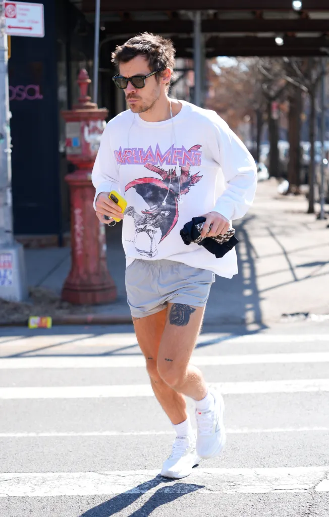 Harry Styles running in a white long-sleeved shirt, gray shorts, and white sneakers, holding a phone and earbuds.