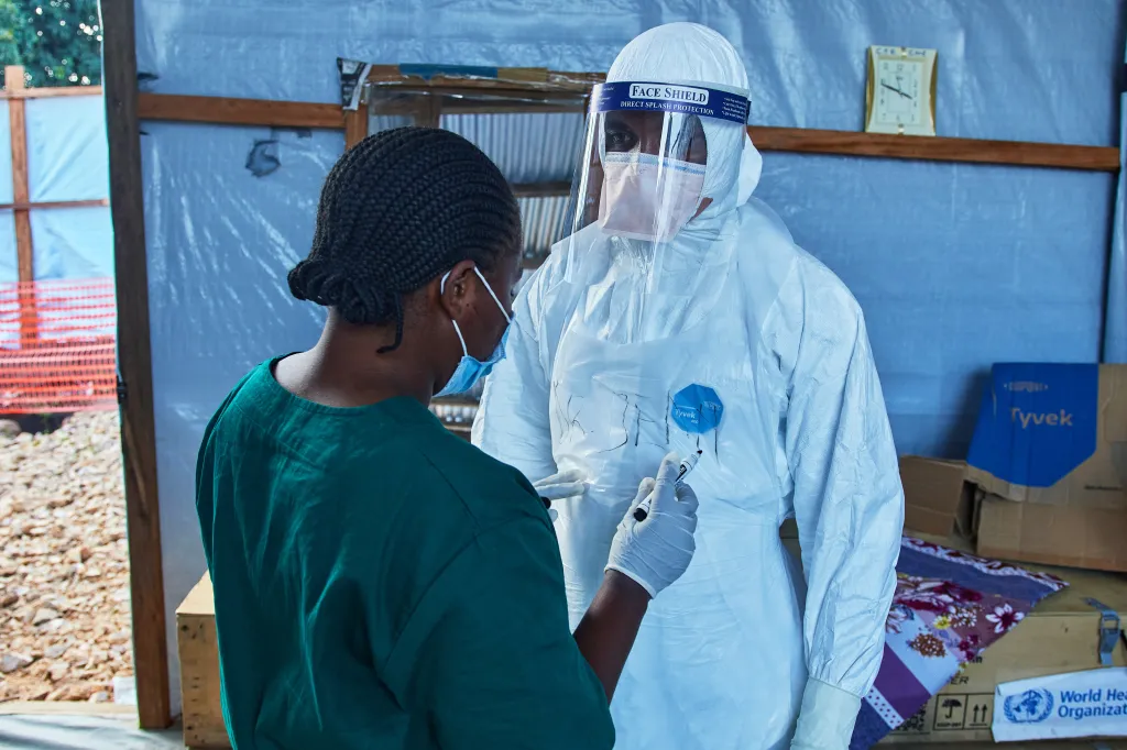 A health worker in a face mask and dark green uniform writing on the full-body protective suit of another health worker.