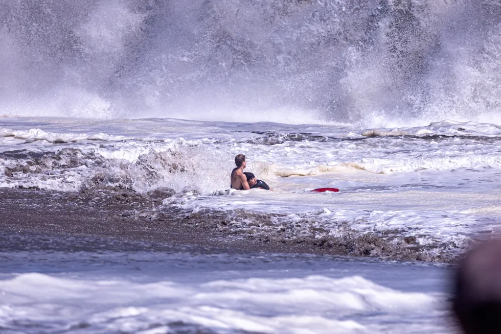 A boy and a lifeguard in rough surf with a large breaking wave in the background.