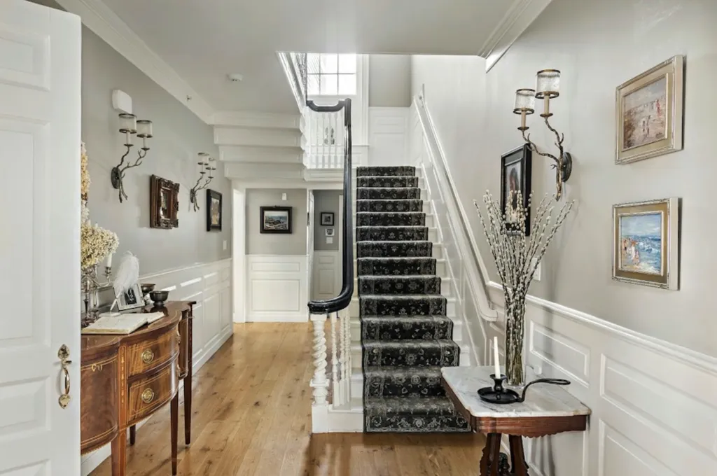 Foyer of a historic Salem home with a grand staircase covered in a dark patterned runner, light wood floors, and decorative wall sconces.