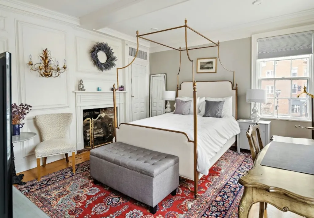 Bedroom with a gold canopy bed, white bedding, red and blue rug, and a fireplace.