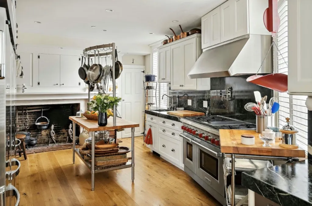 A modern white kitchen with wood floors, white cabinets, stainless steel appliances, and a historic brick fireplace with hanging pots.