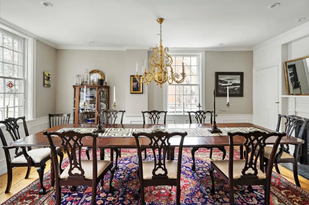A dining room with a long wooden table, dark wooden chairs with cream-colored cushions, a chandelier, and a colorful rug on a light wood floor.