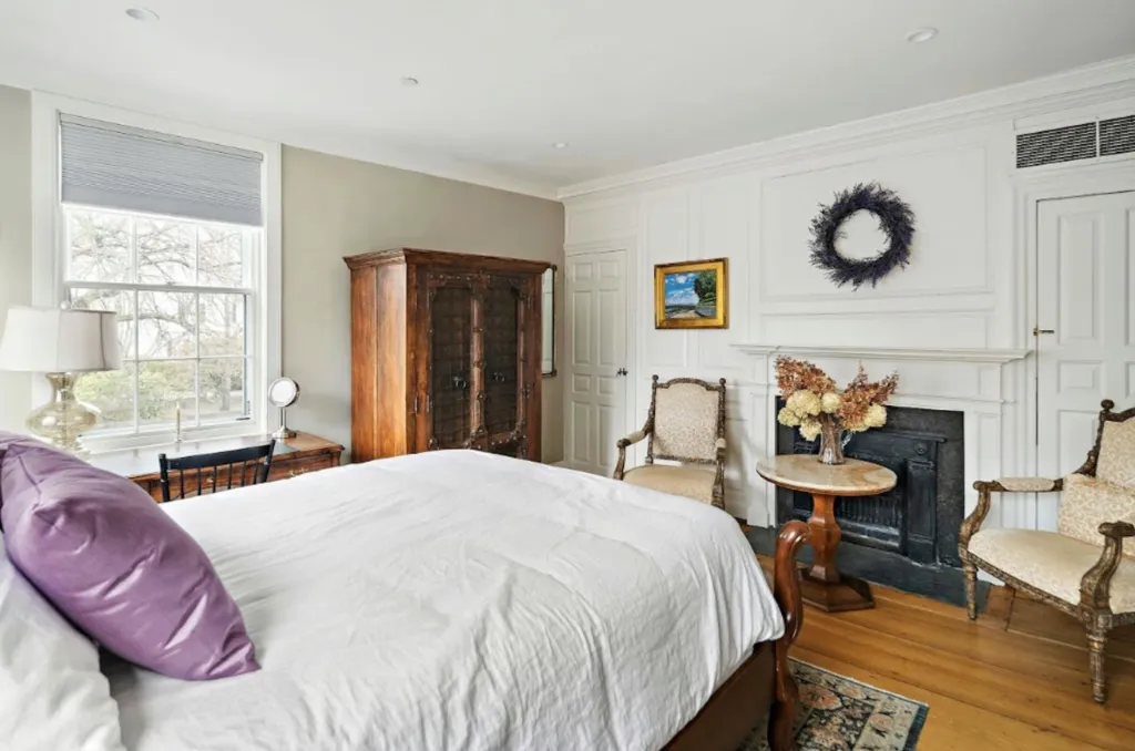 A historic bedroom with a white bed, purple pillow, a fireplace, and antique furniture.