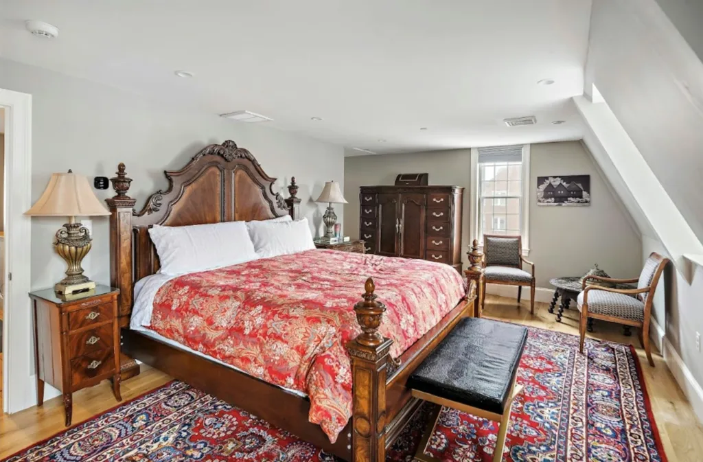 A bedroom in a historic Salem home, featuring a large wooden bed with a red floral duvet, nightstands, lamps, and a red and blue patterned rug.