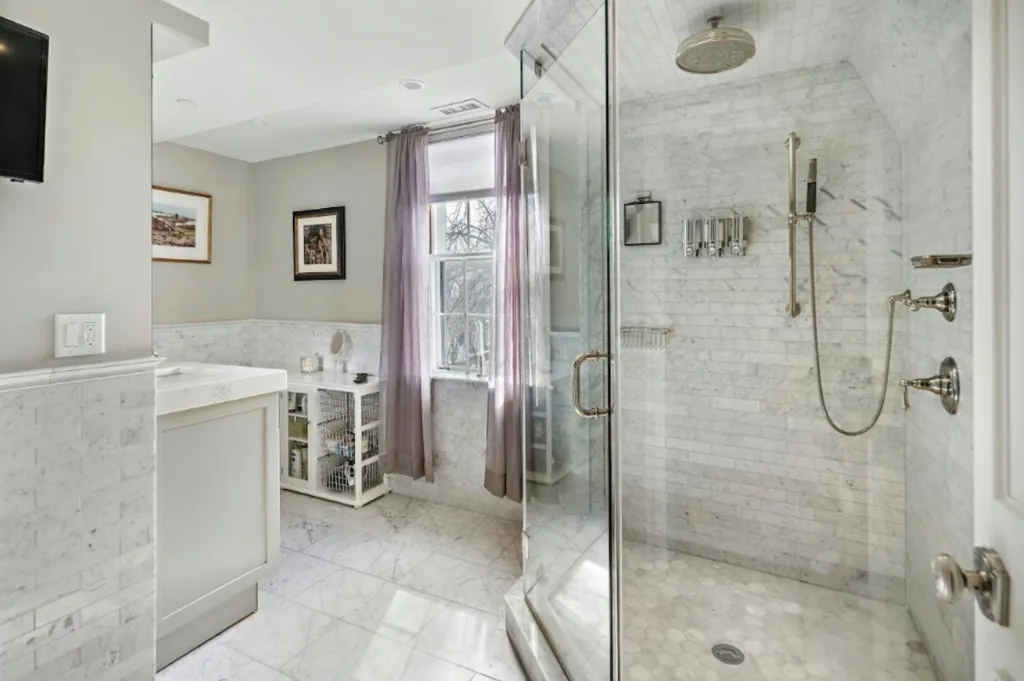A modern bathroom with white marble walls and flooring, a glass shower with a rain shower head, and a window with purple curtains.