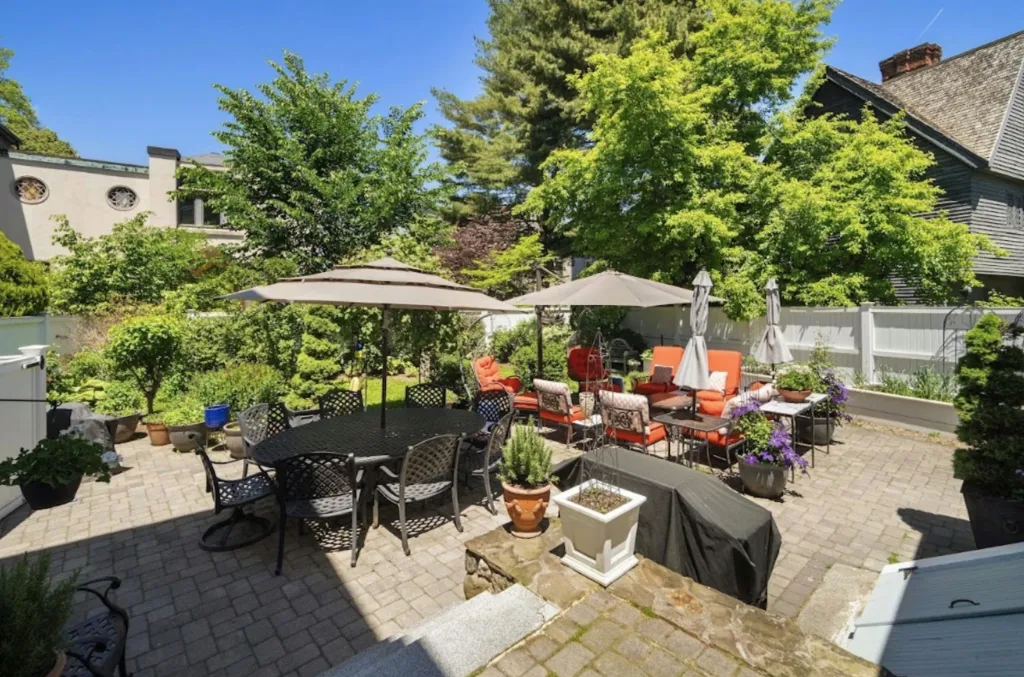 A large brick patio with two seating areas, one for dining and one for lounging, surrounded by lush green foliage and houses in the background.