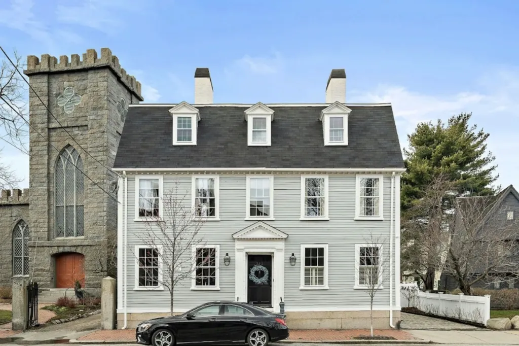 A light gray historic house with a black front door and a dark shingled roof, next to a stone church tower.