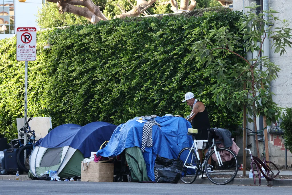A homeless encampment on Hampton Drive with a man standing near blue tarps and tents, with bikes and belongings visible on the street.