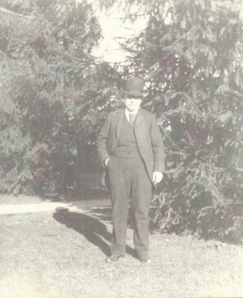 Hugh Alexander Rose II wearing a hat, standing on a lawn in front of evergreens.