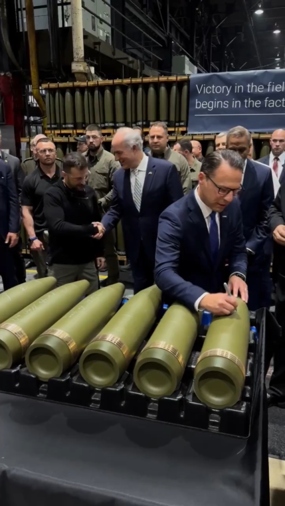 Men in a factory, including Ilhan Omar, signing large green artillery shells.