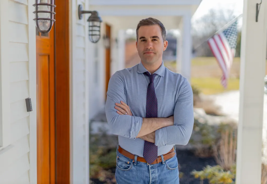 Jordan Wood, candidate for Maine Governor, poses on a porch with an American flag in the background.