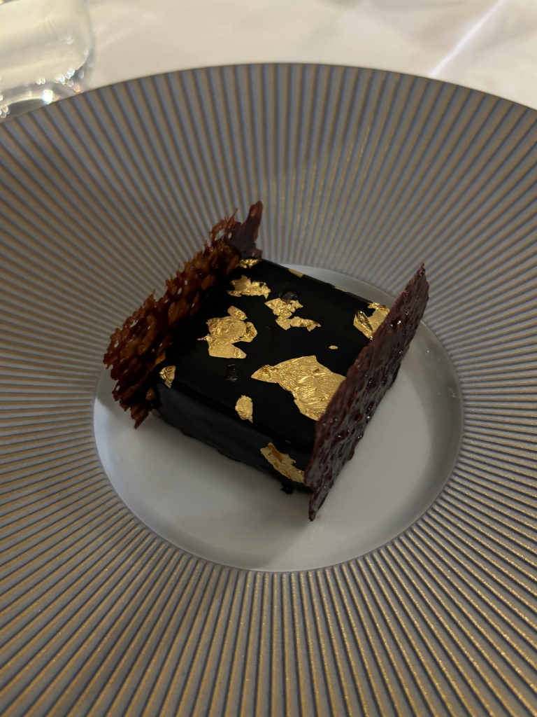 A dark chocolate dessert with gold leafing and a crisp garnish, served on a white plate with golden pinstripes.