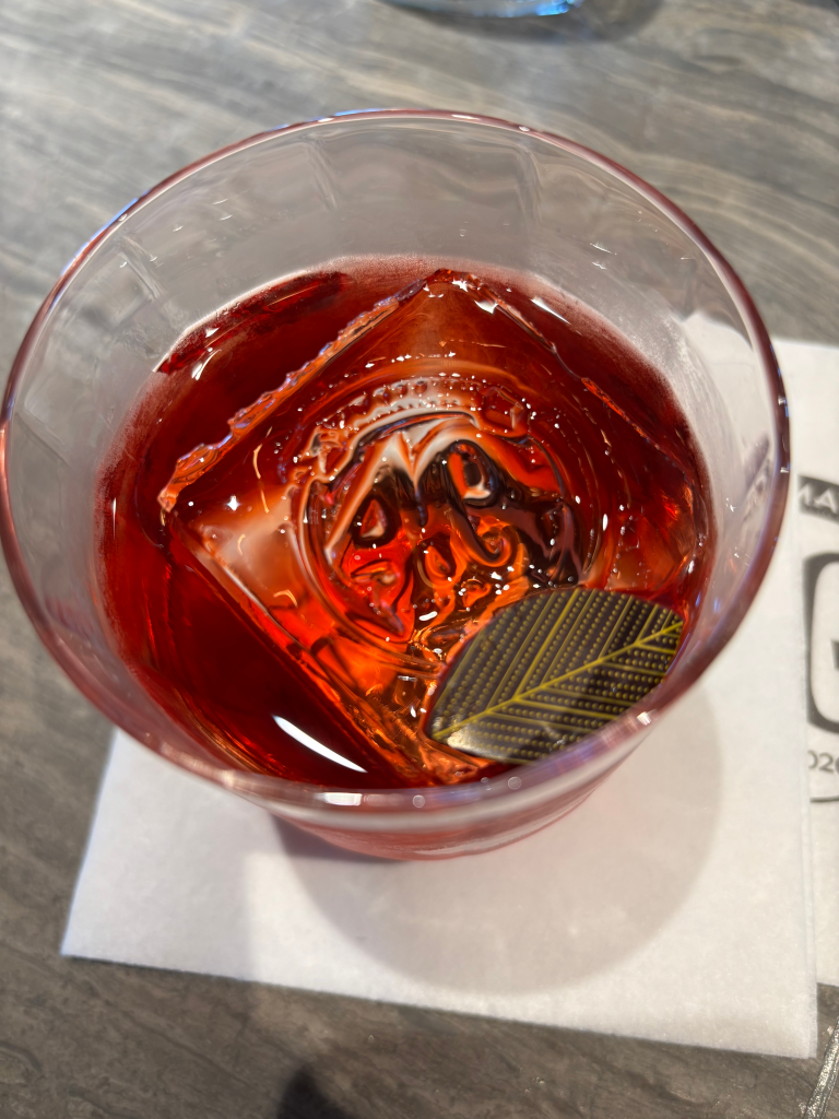A red drink in a clear glass with ice and a dark leaf garnish with yellow veins.