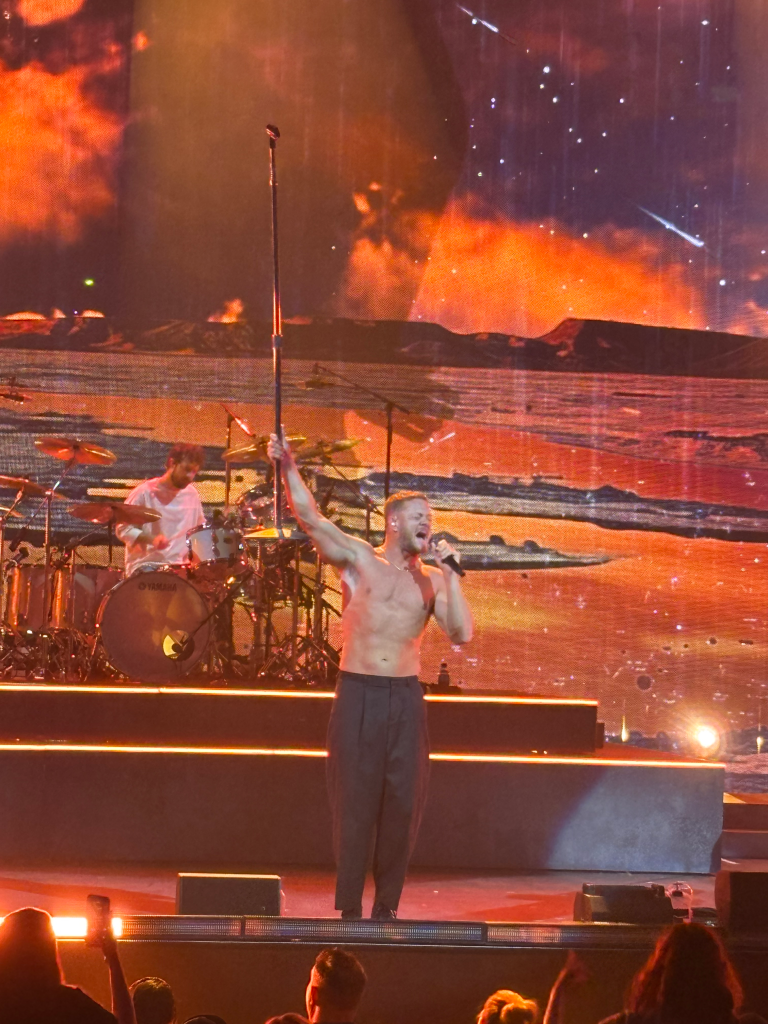 Dan Reynolds of Imagine Dragons performs shirtless with a microphone in hand, lit by orange stage lights.