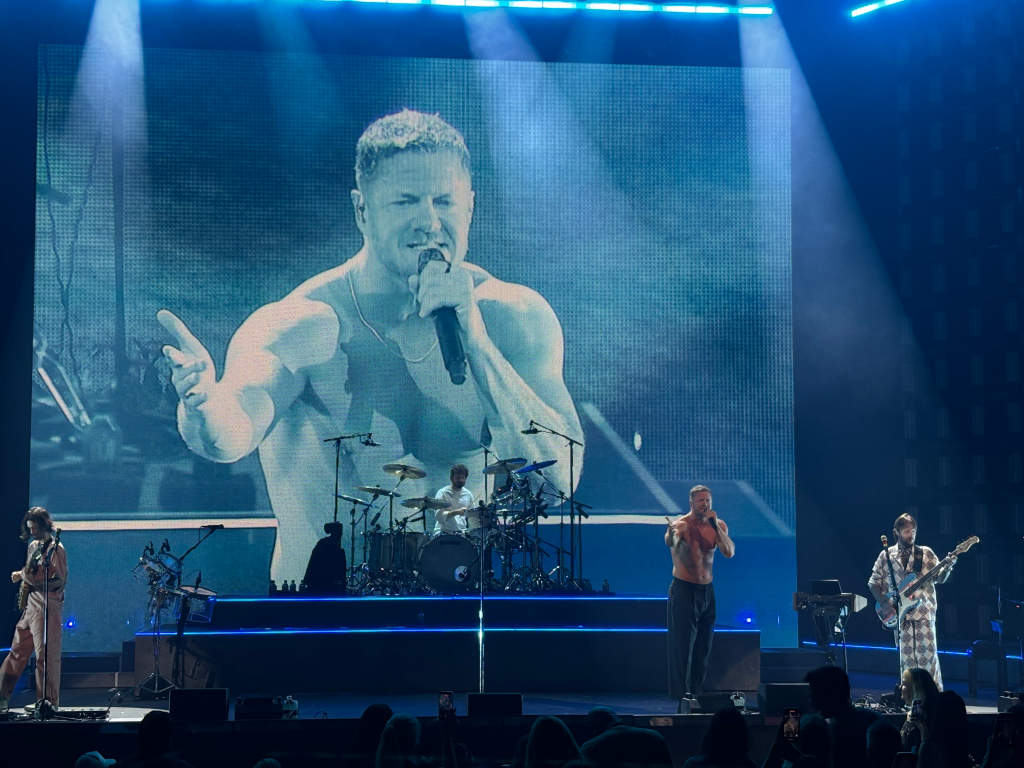 Imagine Dragons performs live with lead singer Dan Reynolds on stage in front of a large screen showing a close-up of him.