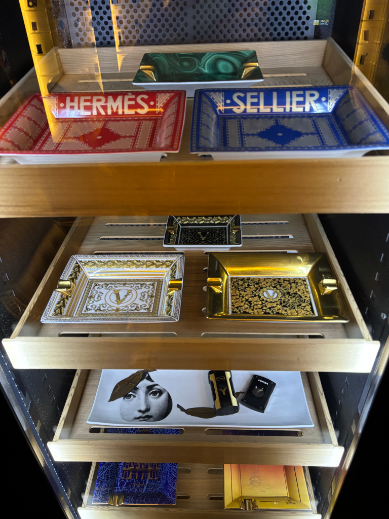 A display case filled with luxury items, including red and blue Hermes ashtrays, several gold and black patterned Versace ashtrays, and a tray with a partial face illustration.