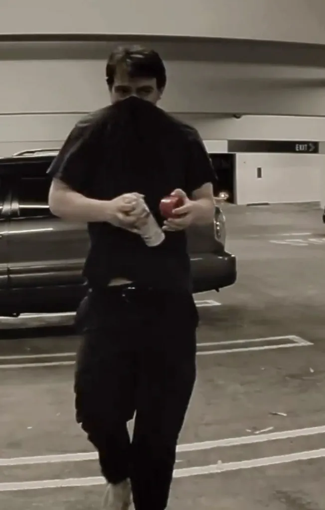 A man covering his face with a black shirt holds a spray paint can and a red object in a parking garage.