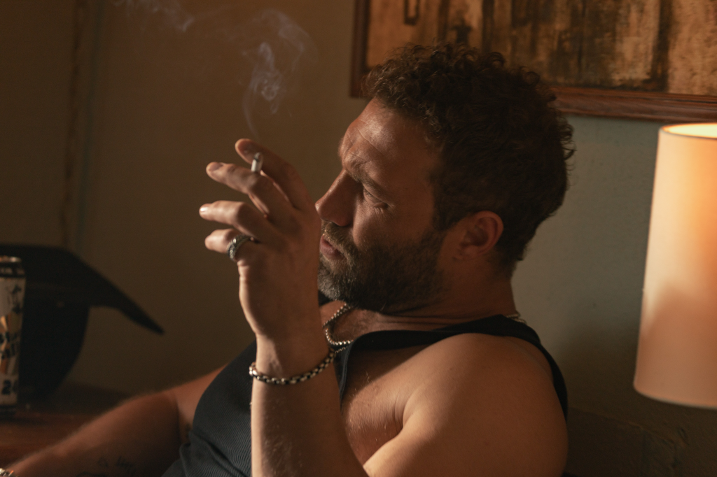 Jai Courtney as Rob-Will, wearing a dark tank top, smoking a cigarette, with a ring and bracelets visible.