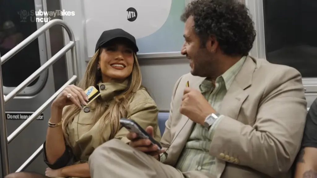 Jennifer Lopez holding a MetroCard and sitting next to a man on a subway.