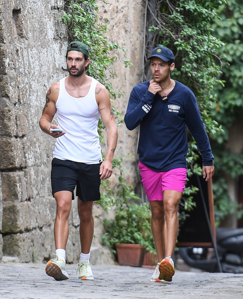 Harry Styles, in a navy blue top and pink shorts and a blue baseball cap, jogging in Bagnoregio, Italy, with his personal trainer Brad Gould, in a white tank top and black shorts.