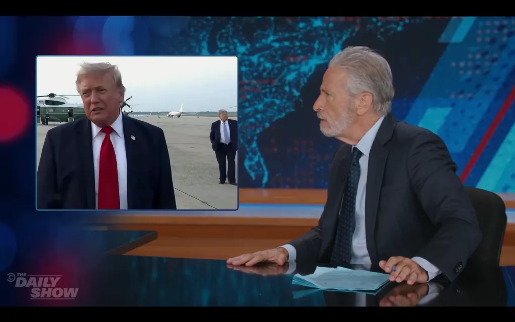 Jon Stewart looks surprised at a screen showing Donald Trump speaking with a man resembling him in the background.