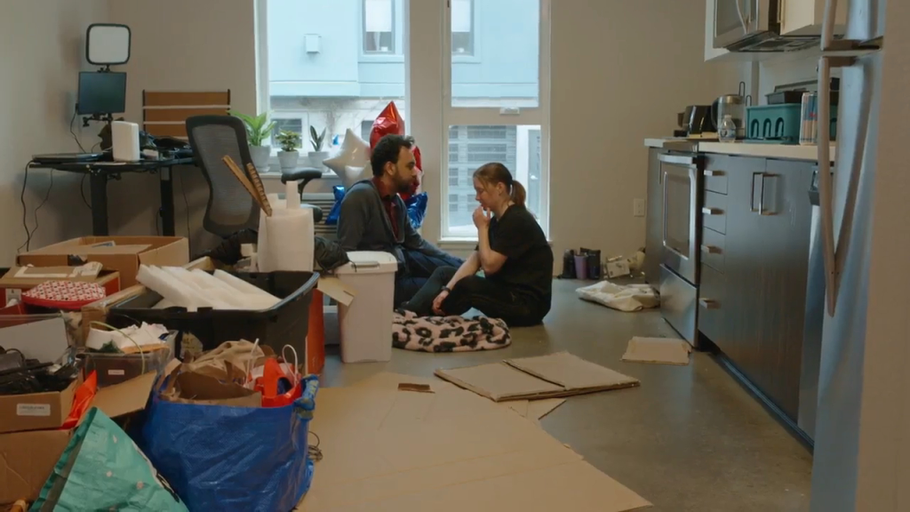 Josh and Catie on the floor talking in a messy room.