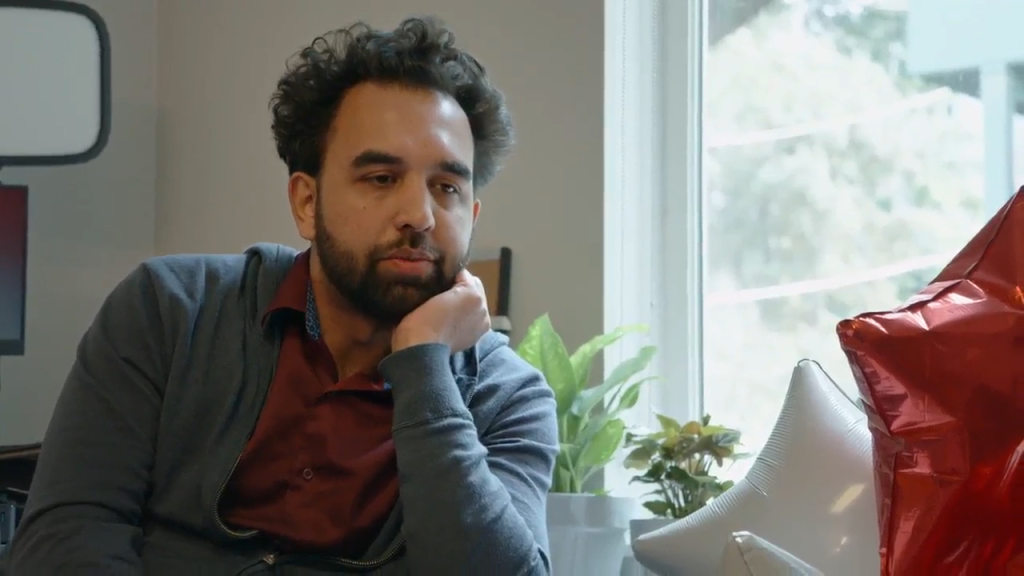 A man with dark, curly hair and a beard rests his chin on his hand, looking pensive.