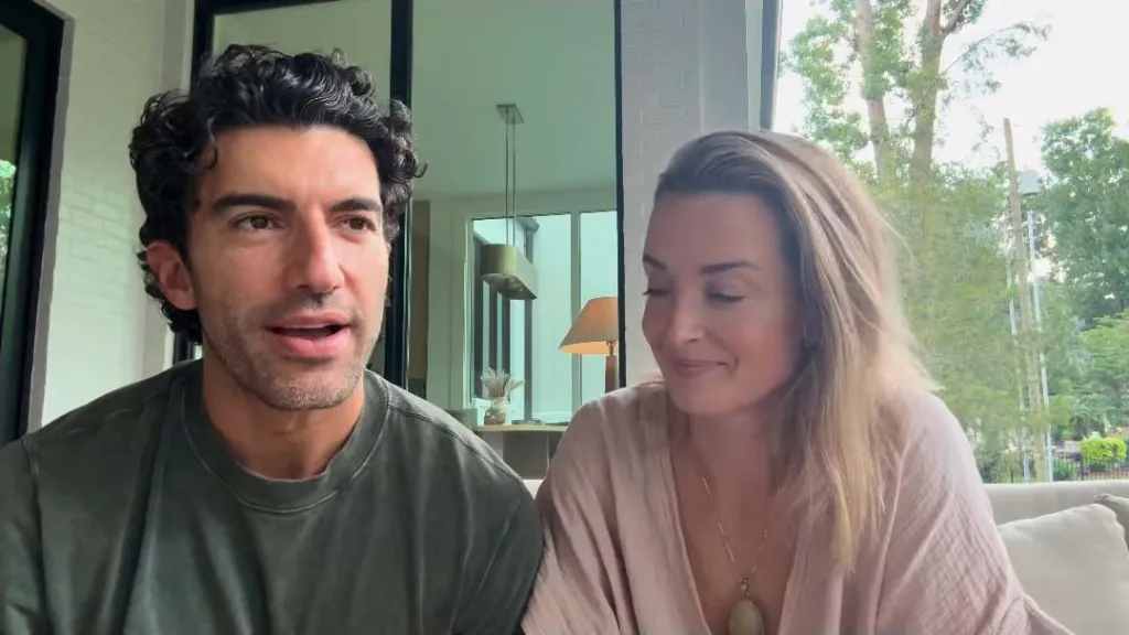 Justin Baldoni and Emily Baldoni sitting together, with Justin speaking to the camera and Emily looking at him and smiling.