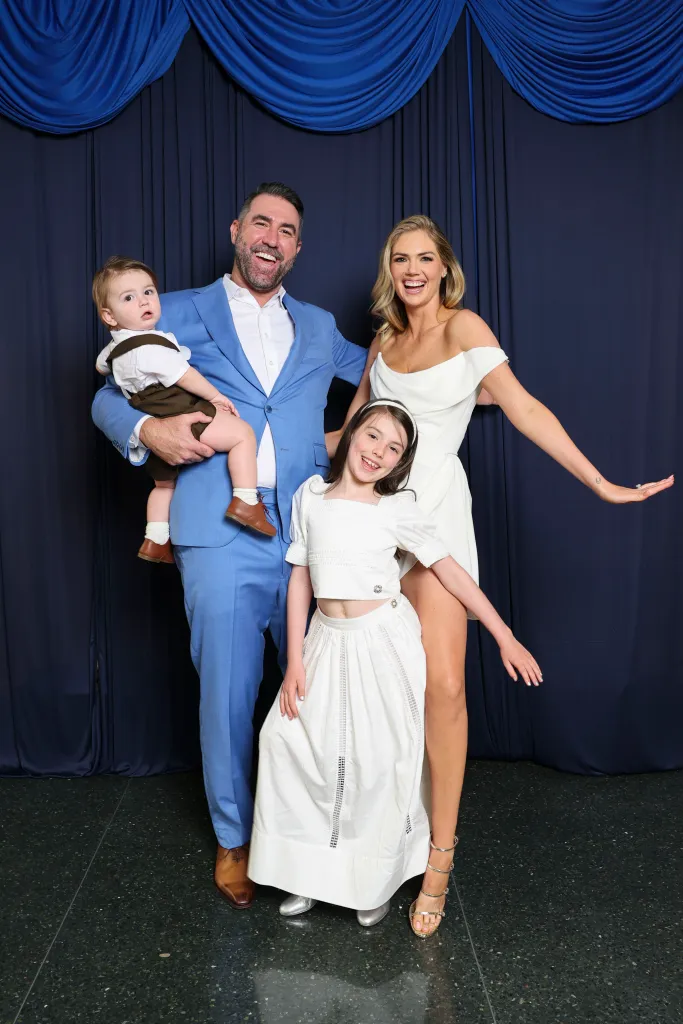Justin Verlander poses with his wife Kate Upton, a young girl, and a baby boy against a blue curtain backdrop.