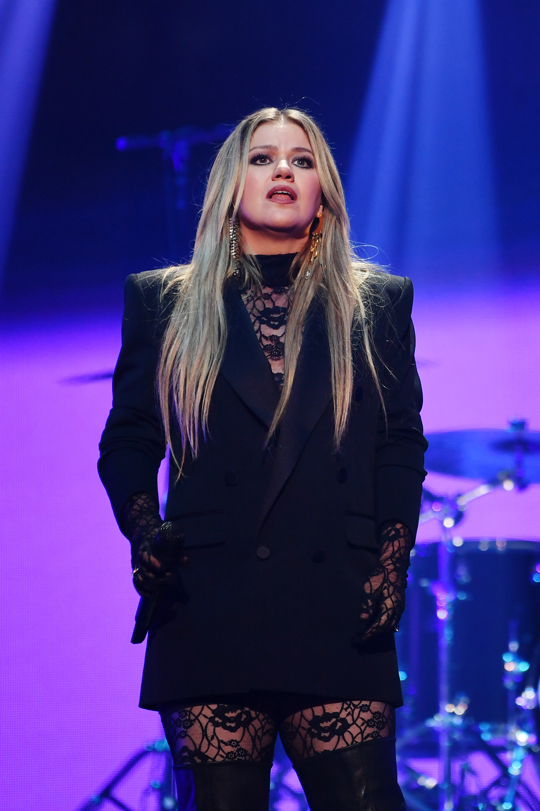 Kelly Clarkson performing onstage, wearing a black double-breasted blazer, black lace gloves, and lace thigh-high stockings.