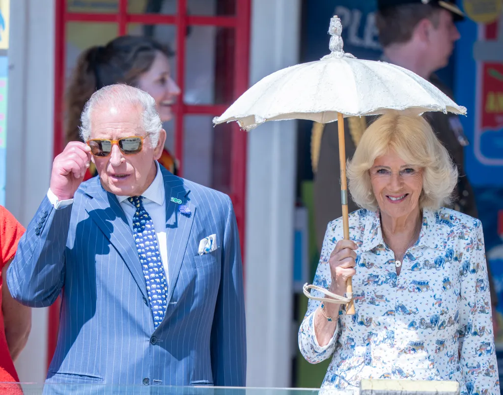 King Charles III, in a blue pin-striped suit, blue tie and white shirt and sunglasses, and Queen Camilla, in a white dress with blue floral pattern, holding an umbrella at Penguin Beach during a visit to London Zoo.