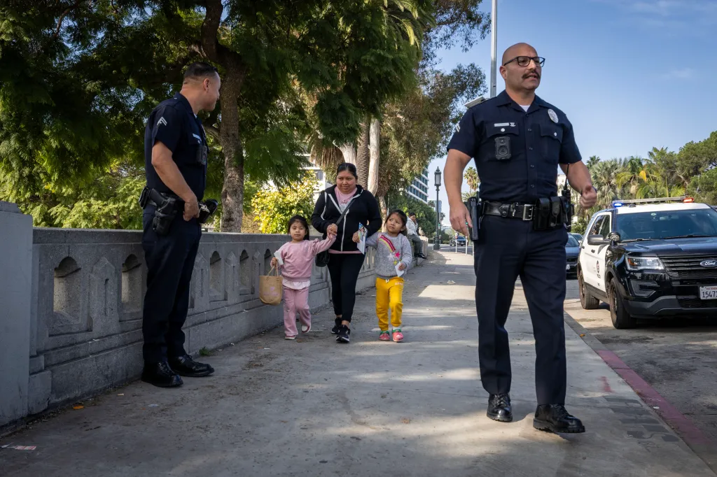 Two police officers stand on a sidewalk while a woman and two children walk past them.
