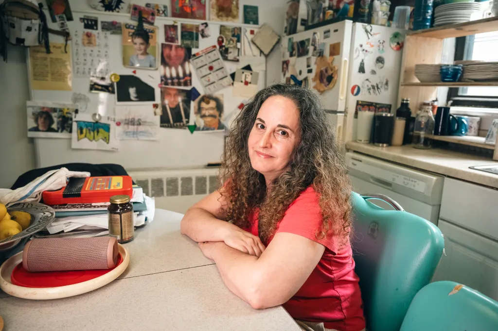 Lara Kossover in a pink shirt sits at a table in a aqua chair with her arms folded on the table.