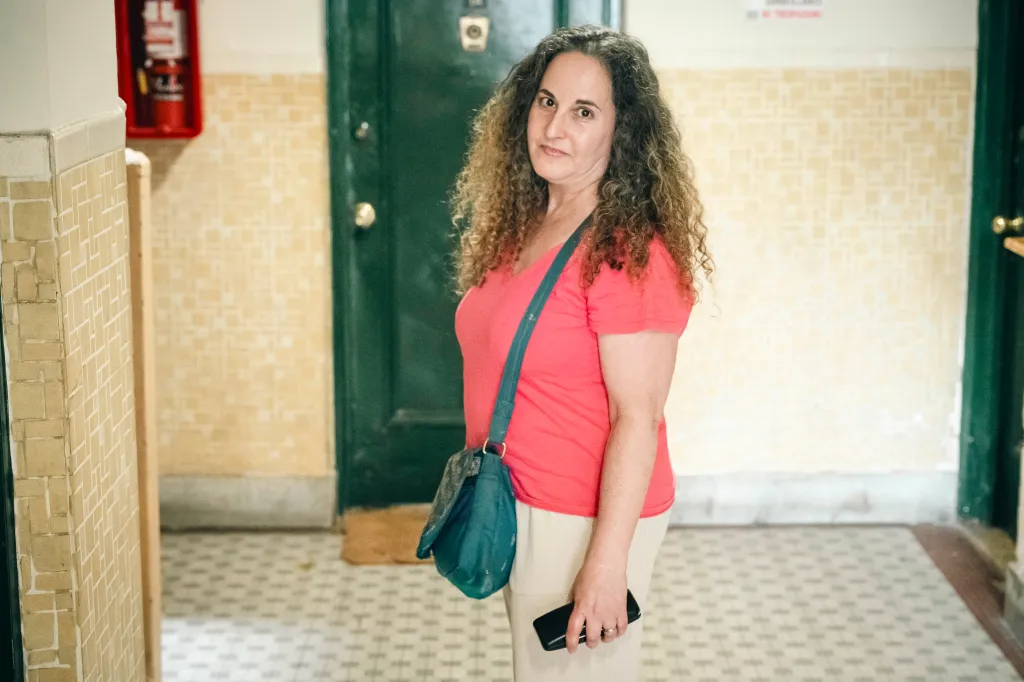 Lara Kossover dressed in a pink shirt, tan pants and a green crossbody bag stands in the hallway of a building.
