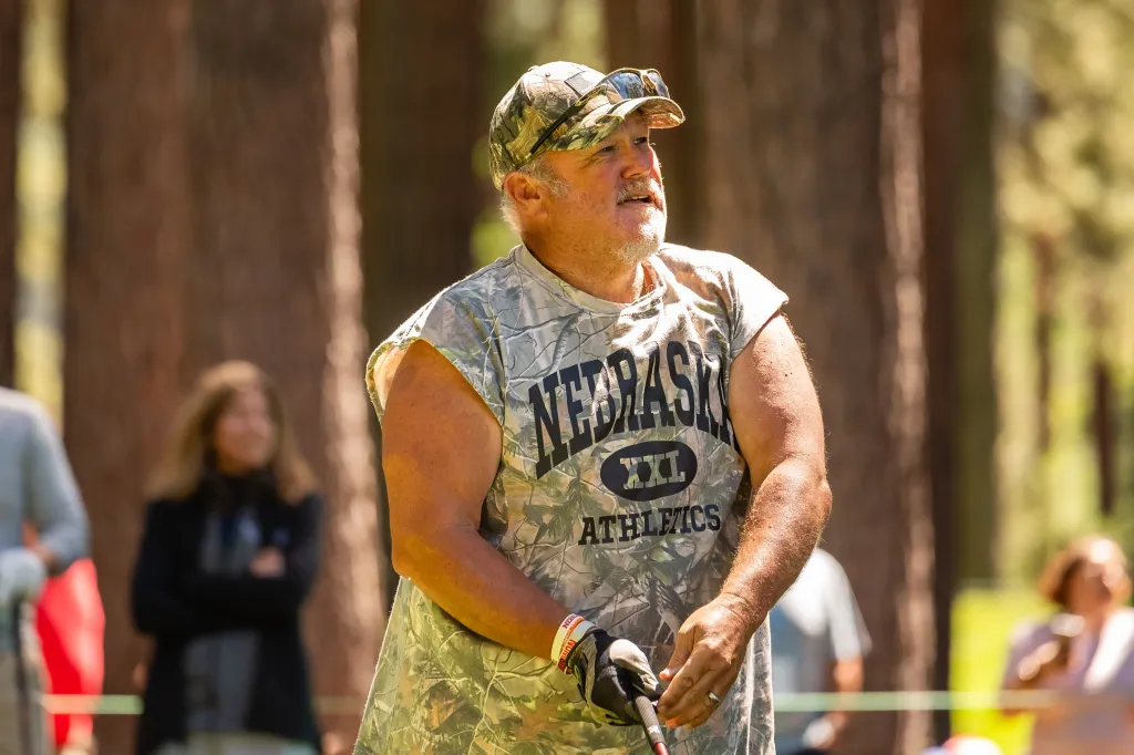 Larry the Cable Guy wearing a camouflage shirt and hat, playing golf.