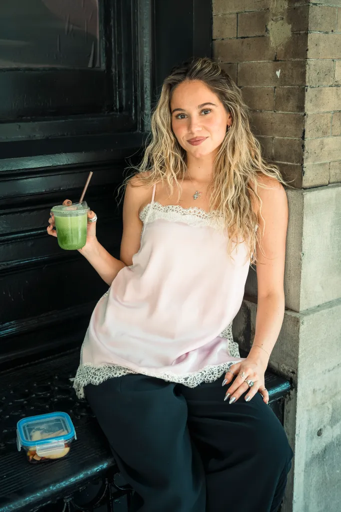 Lauren Holaday in a pink lace-trimmed top and black pants, holding a green drink and sitting on outdoor steps in Nolita.