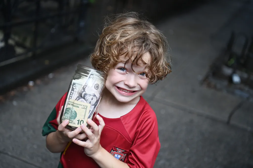 A smiling child in a red shirt holds a jar full of money.