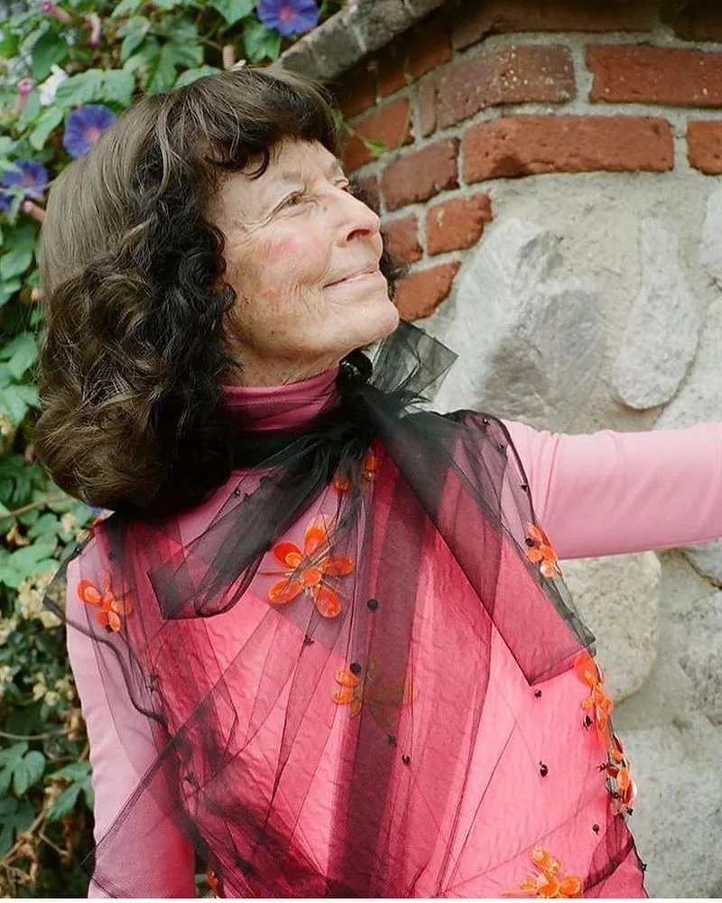 Linda Perhacs, a woman with dark hair, looking up and to the right, wearing a pink top and a sheer black shawl with orange floral embellishments.