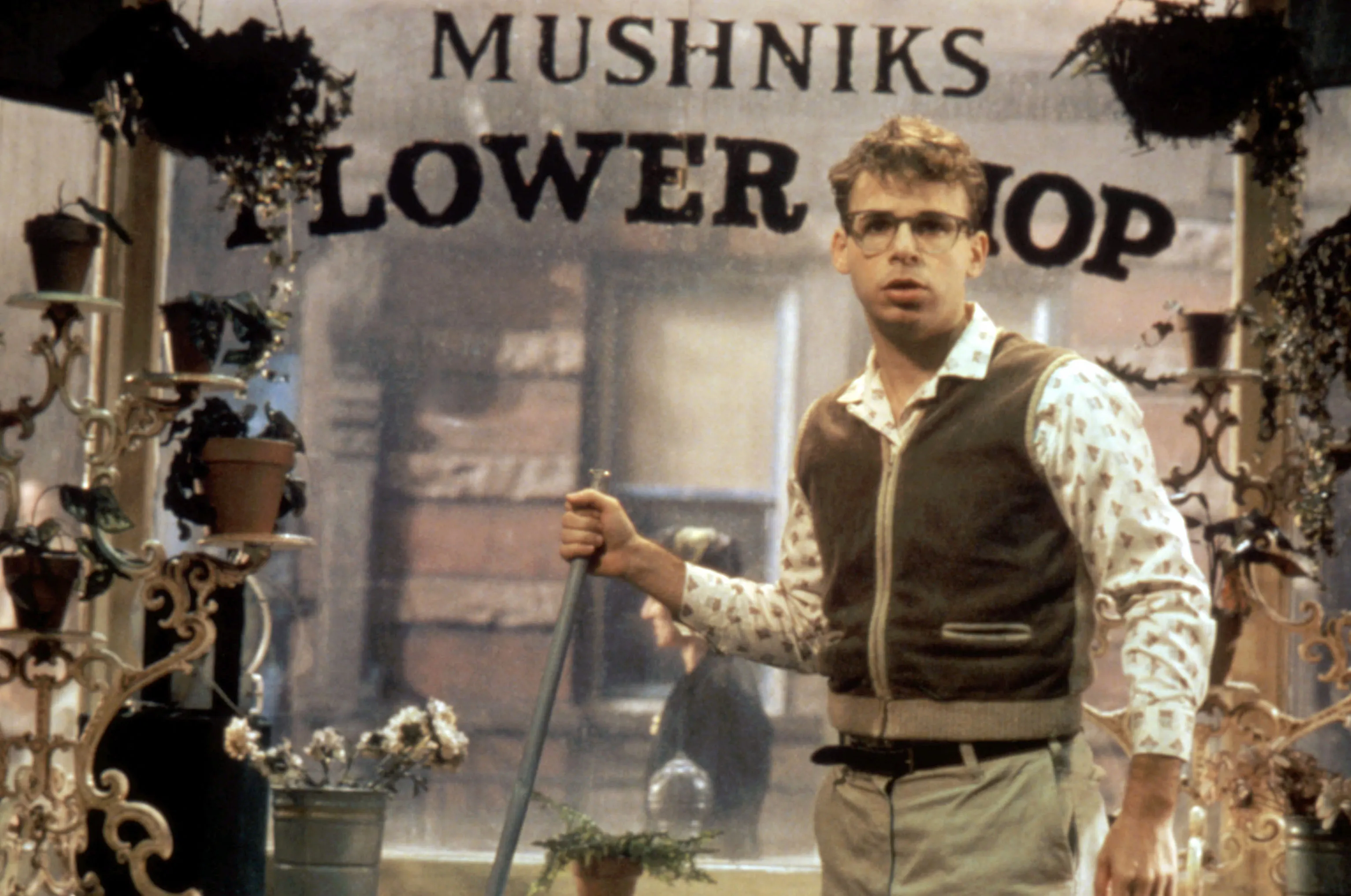 Rick Moranis in character as Seymour, a nerd with eyeglasses, a vest, and a patterned shirt, holding a broom in front of Mushnik's Flower Shop in the film Little Shop of Horrors.