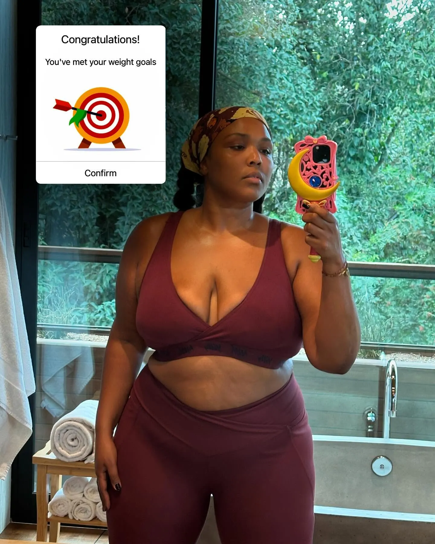 Lizzo poses for a mirror selfie in a maroon sports bra and leggings, with a pop-up congratulating her for meeting her weight goals.