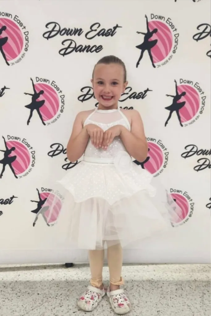 Calli Toler, a 6-year-old girl, smiling in a white dance costume in front of a 
