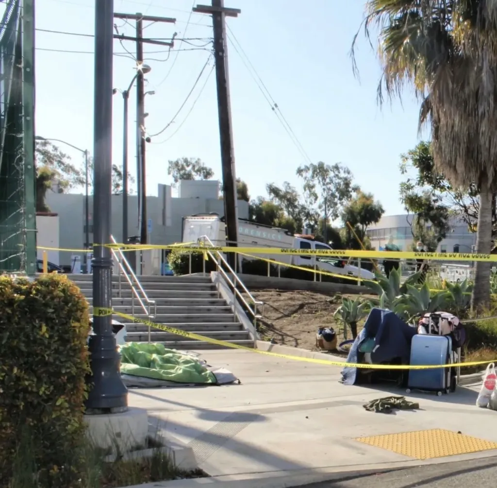Yellow police tape blocks off a public staircase, with a green sleeping bag and a blue suitcase with belongings visible on the ground.