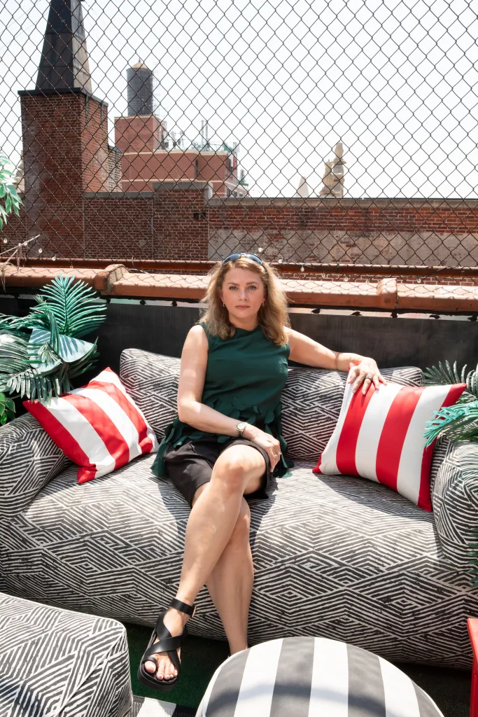 Manhattan real estate publicist Kelly Kreth sits in a green dress on a light couch with red and white striped pillows on a Manhattan rooftop.