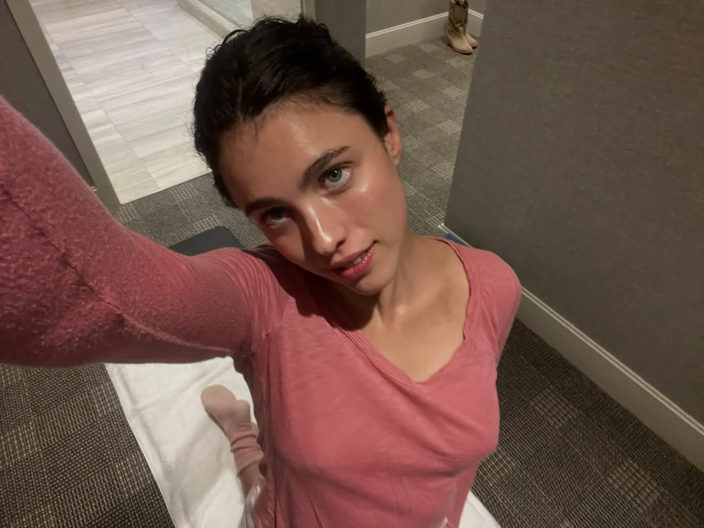 Margaret Qualley takes a selfie while kneeling on a white towel on a gray carpet.