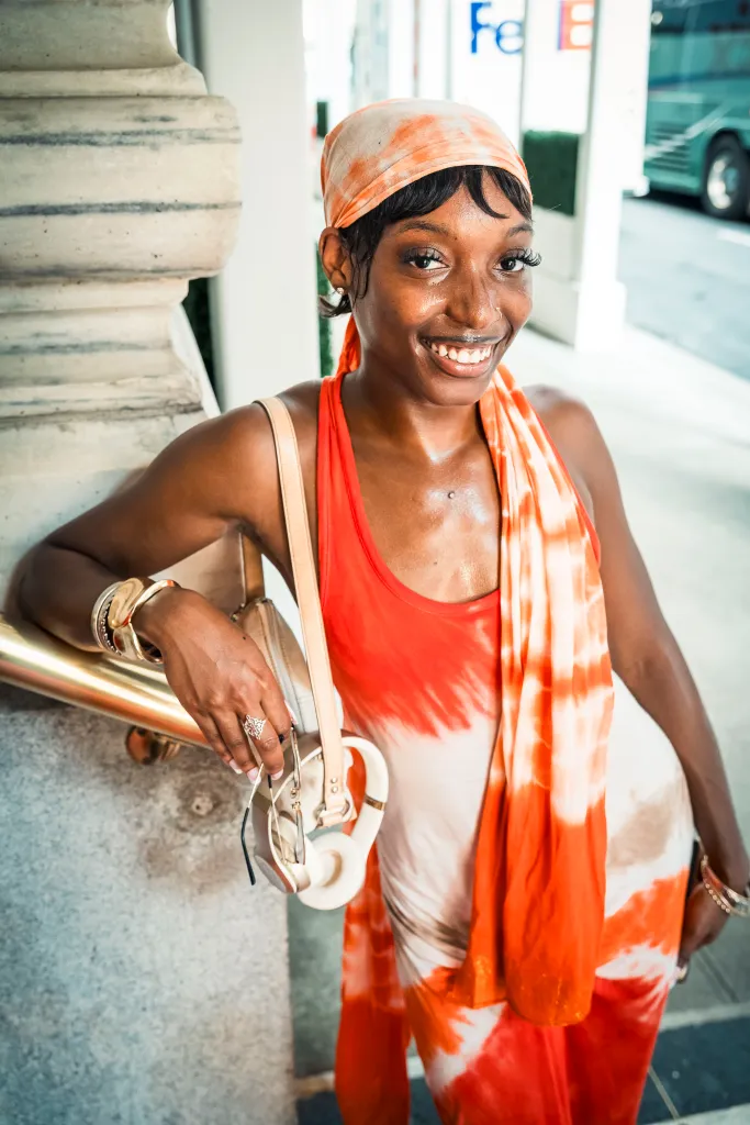 A woman in an orange and white tie-dye dress with an orange headscarf smiles at the camera, leaning against a railing.