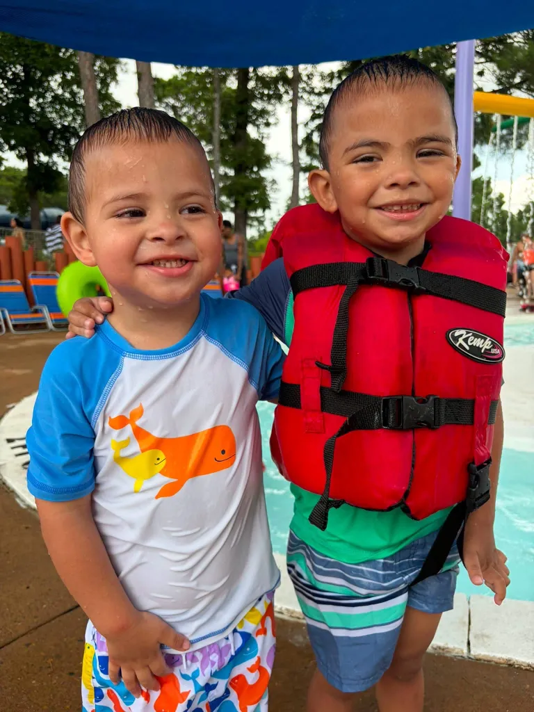 Two young boys, both wet, smile at the camera while standing by a pool.