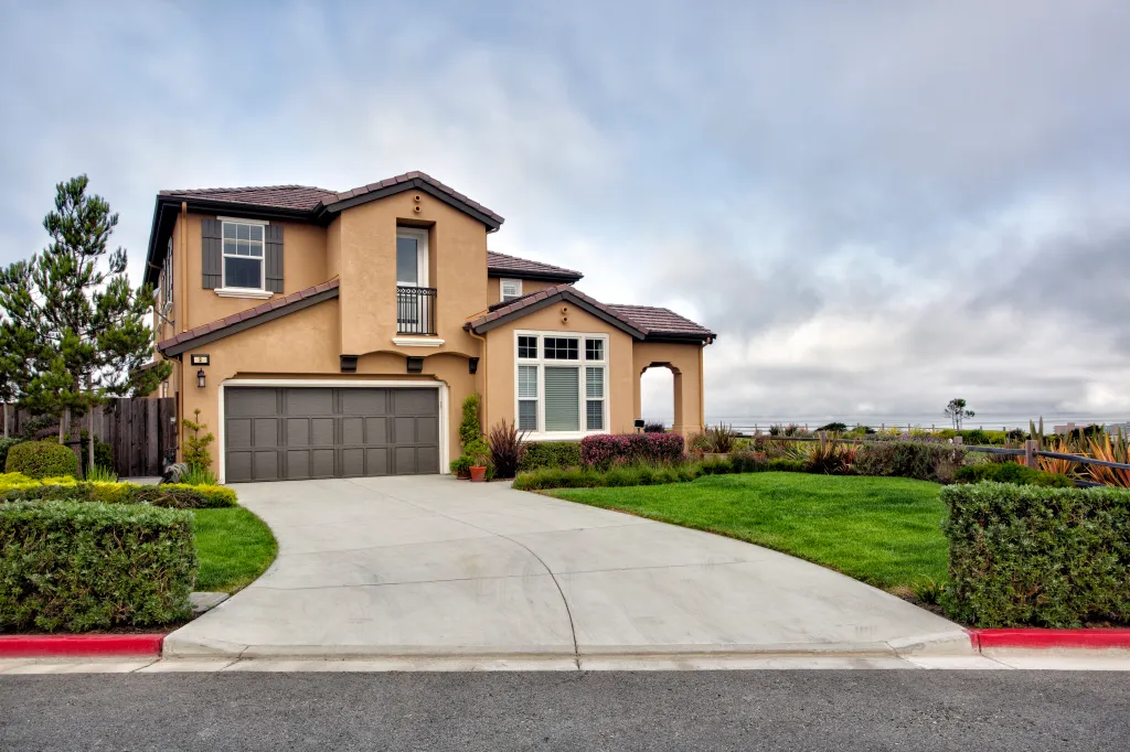 Modern stucco house with a driveway and lush landscaping under an overcast sky.