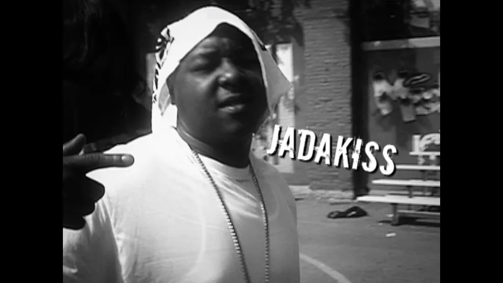 Jadakiss, a Black man in a durag and white t-shirt, stands next to