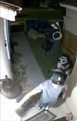 Security camera footage of a man in a t-shirt, shorts, sandals, and a baseball cap standing on a porch.